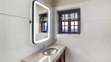 A bathroom with white tiled walls, a large mirror with a light, a sink, and a small window with a view.
