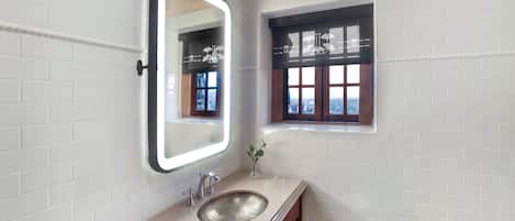 Casa de banho com paredes em azulejo branco, um espelho grande com luz, uma pia e uma pequena janela com vista.