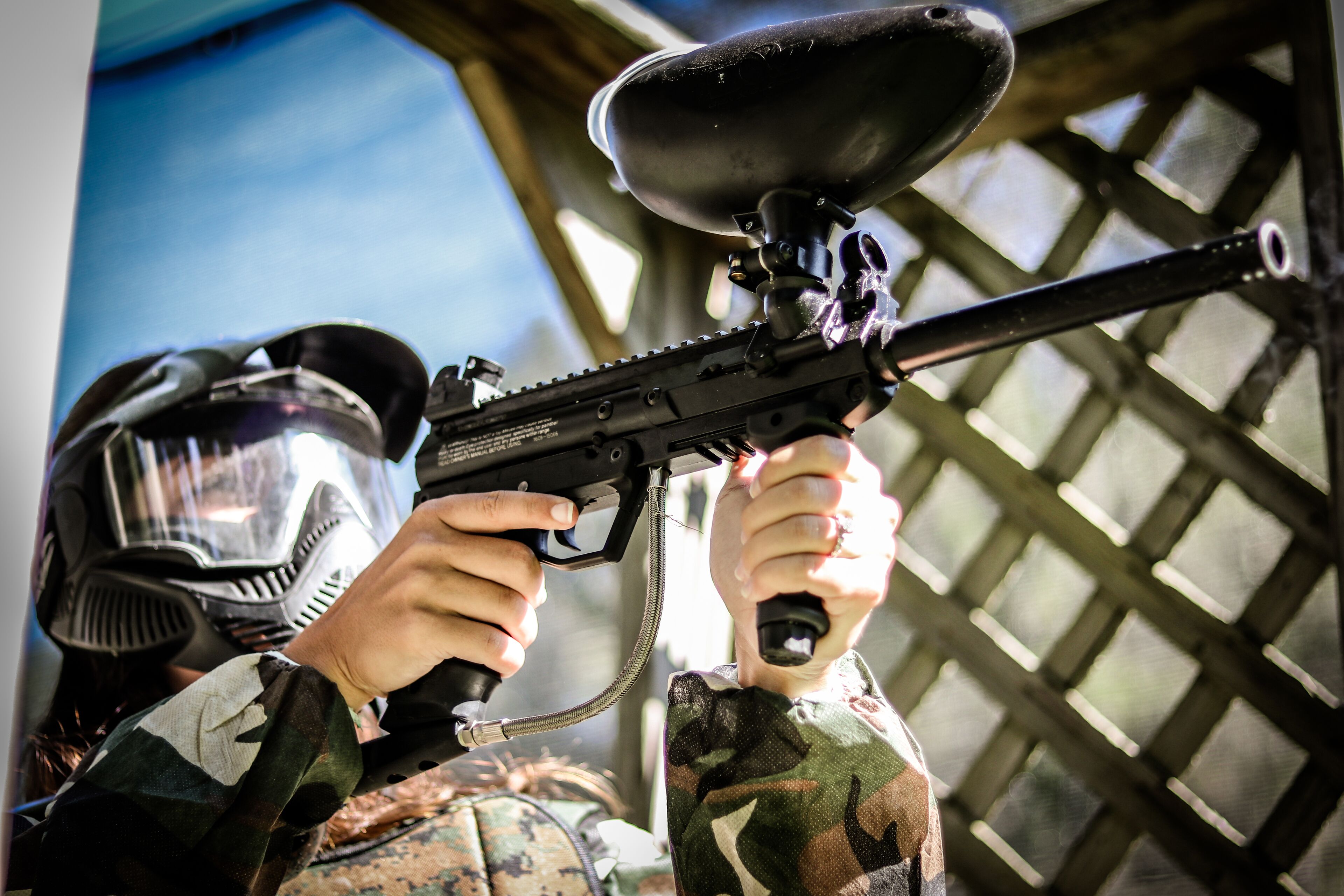 A person holding a paintball gun with a hopper attached, wearing a helmet and camouflage clothing.