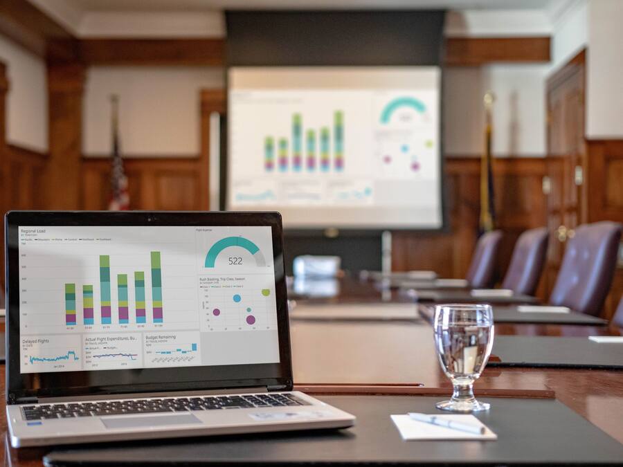 Una laptop mostrando una presentación de negocios con gráficos e informes, un vaso de agua y un salón de conferencias.
