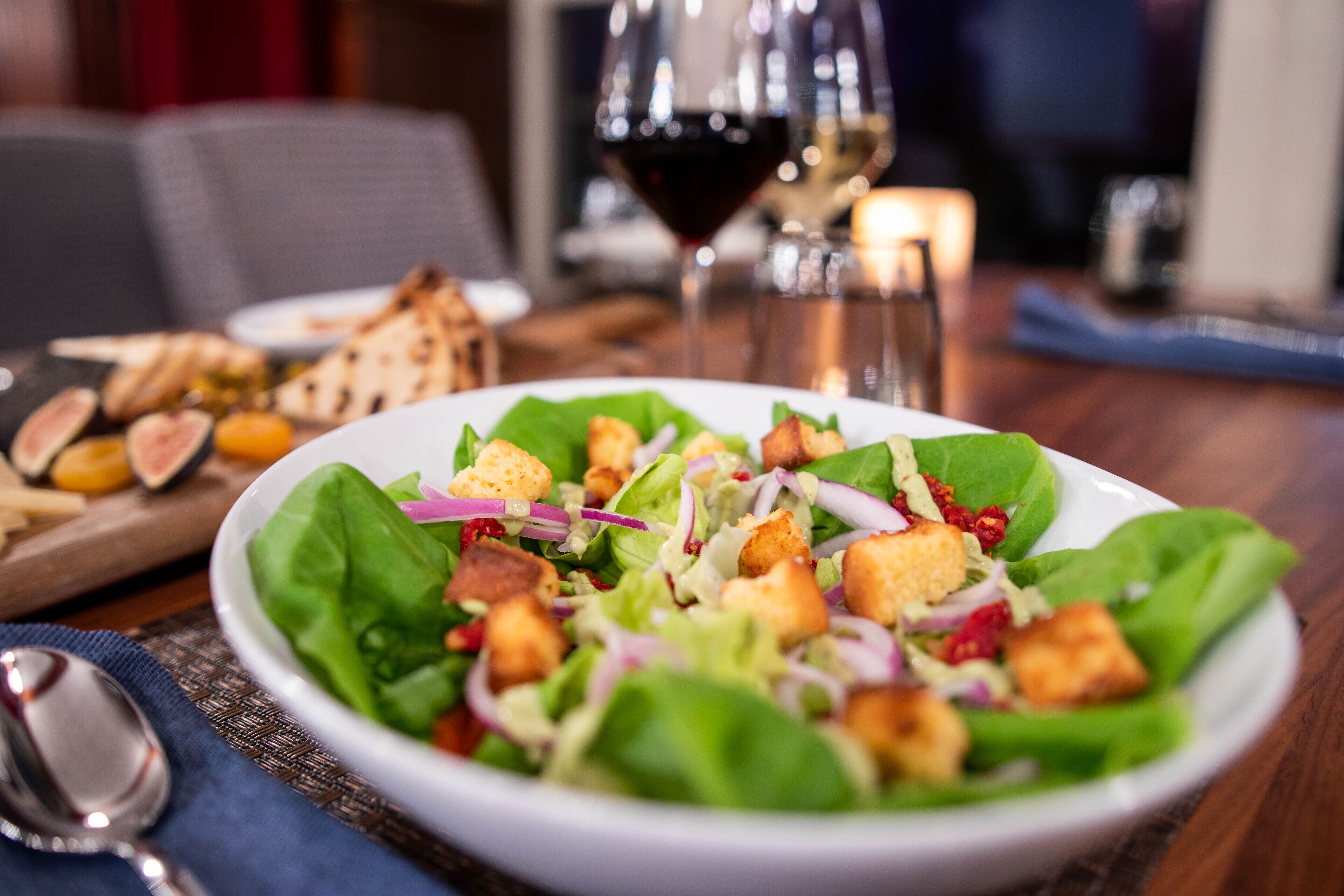 A plate of salad with croutons and red onion, accompanied by a glass of red wine and a candle on a table.