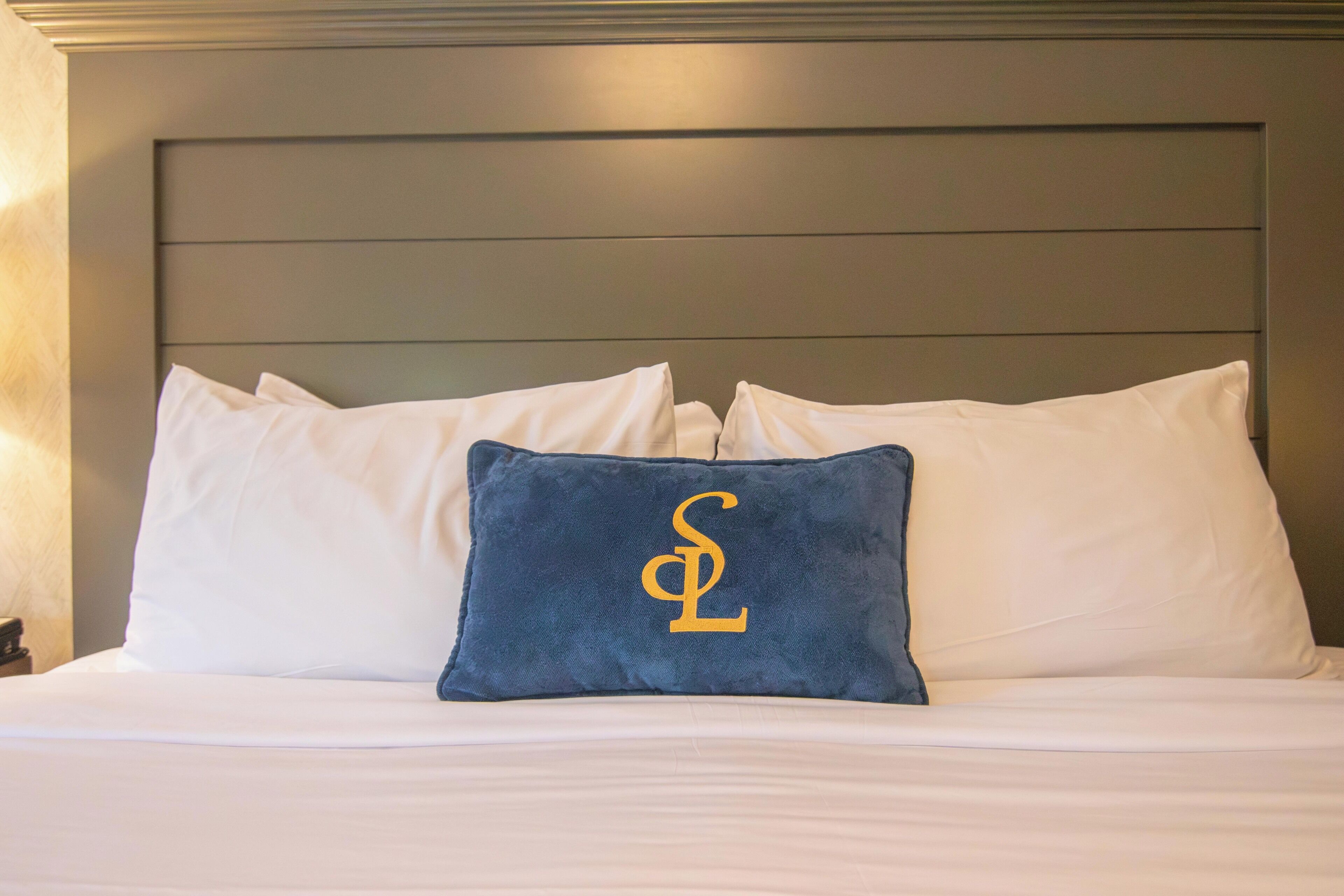 A bed with white bedding and a single blue pillow with a gold letter 'S' on a dark headboard.