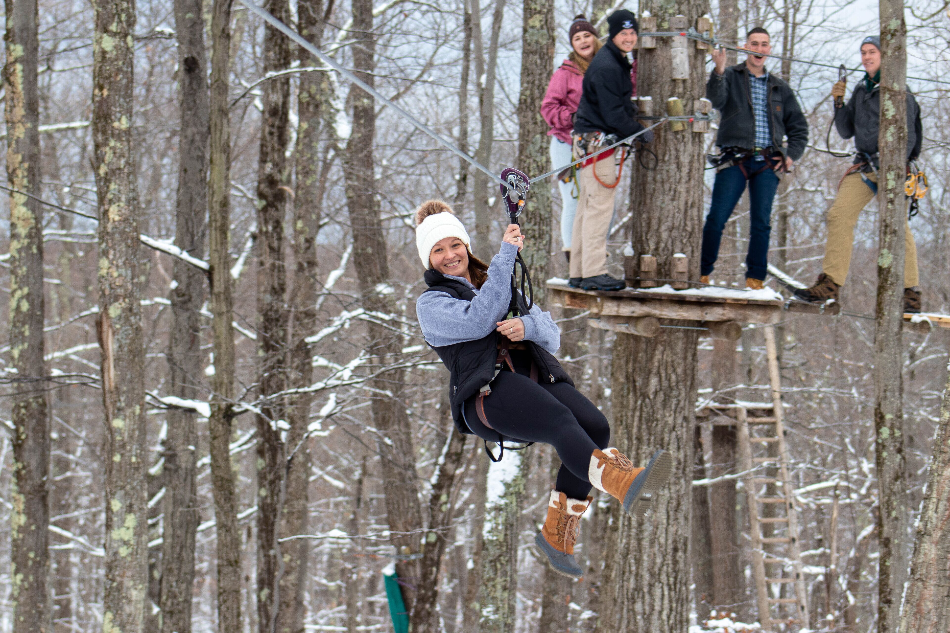 A person zip-lining through a forest with bare trees and snow on the ground.