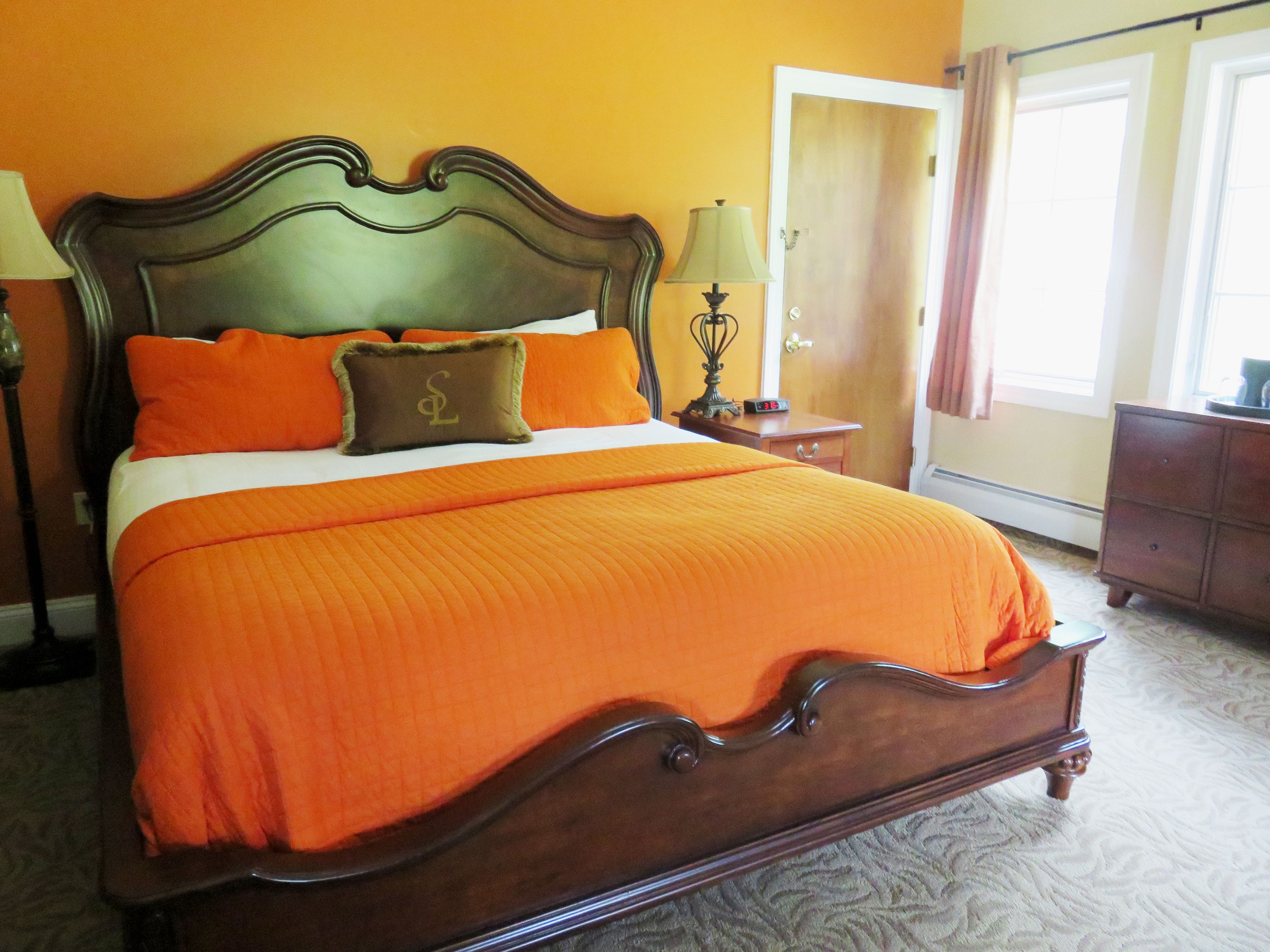 A bed with an orange bedspread and pillows, a wooden headboard, a nightstand with a lamp, and a dresser.