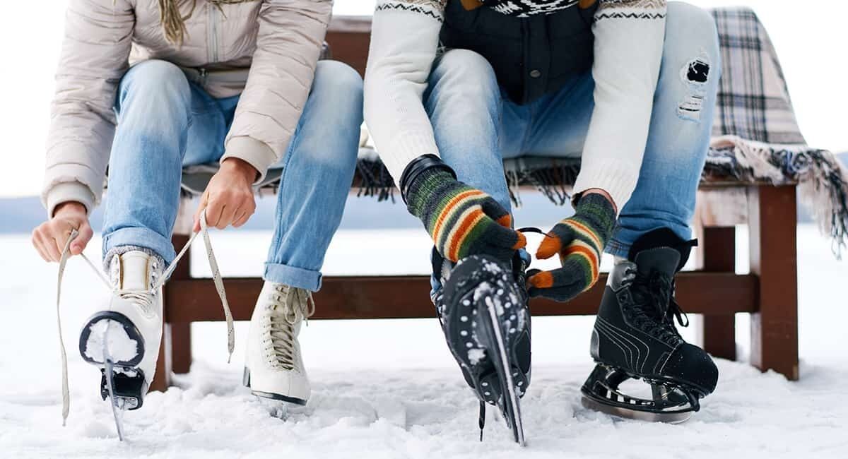 Two people sitting on a bench in a snowy outdoor area, wearing winter clothing and ice skates.