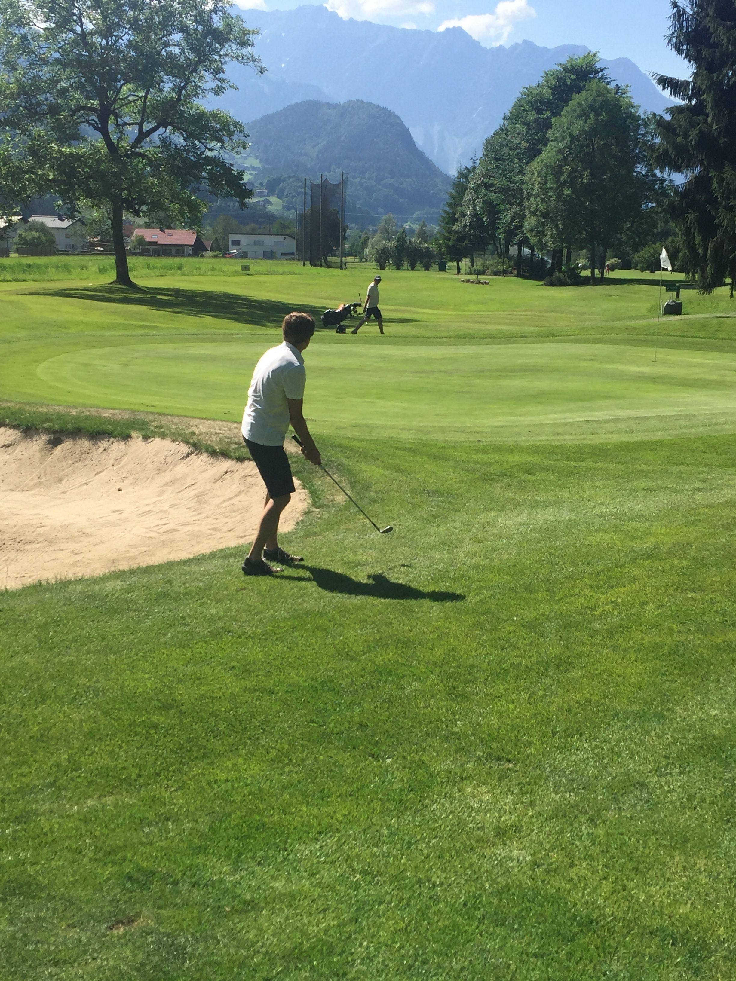 Ein Golfplatz mit einer Person, die sich zum Putten vorbereitet, umgeben von Bäumen und Bergen im Hintergrund.