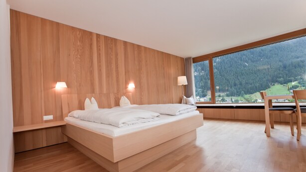 Un dormitorio moderno con cabecera de madera, una cama grande, lámparas de noche y vistas a un paisaje verde a través de un amplio ventanal.