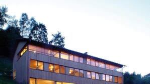 Un edificio moderno de dos plantas con ventanas grandes y techo inclinado, rodeado de árboles.