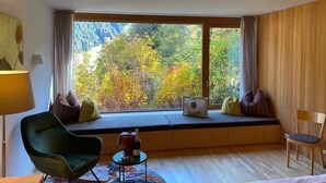 Una habitación con un ventanal grande, una silla verde, una silla de madera y una mesita con una botella y un vaso.