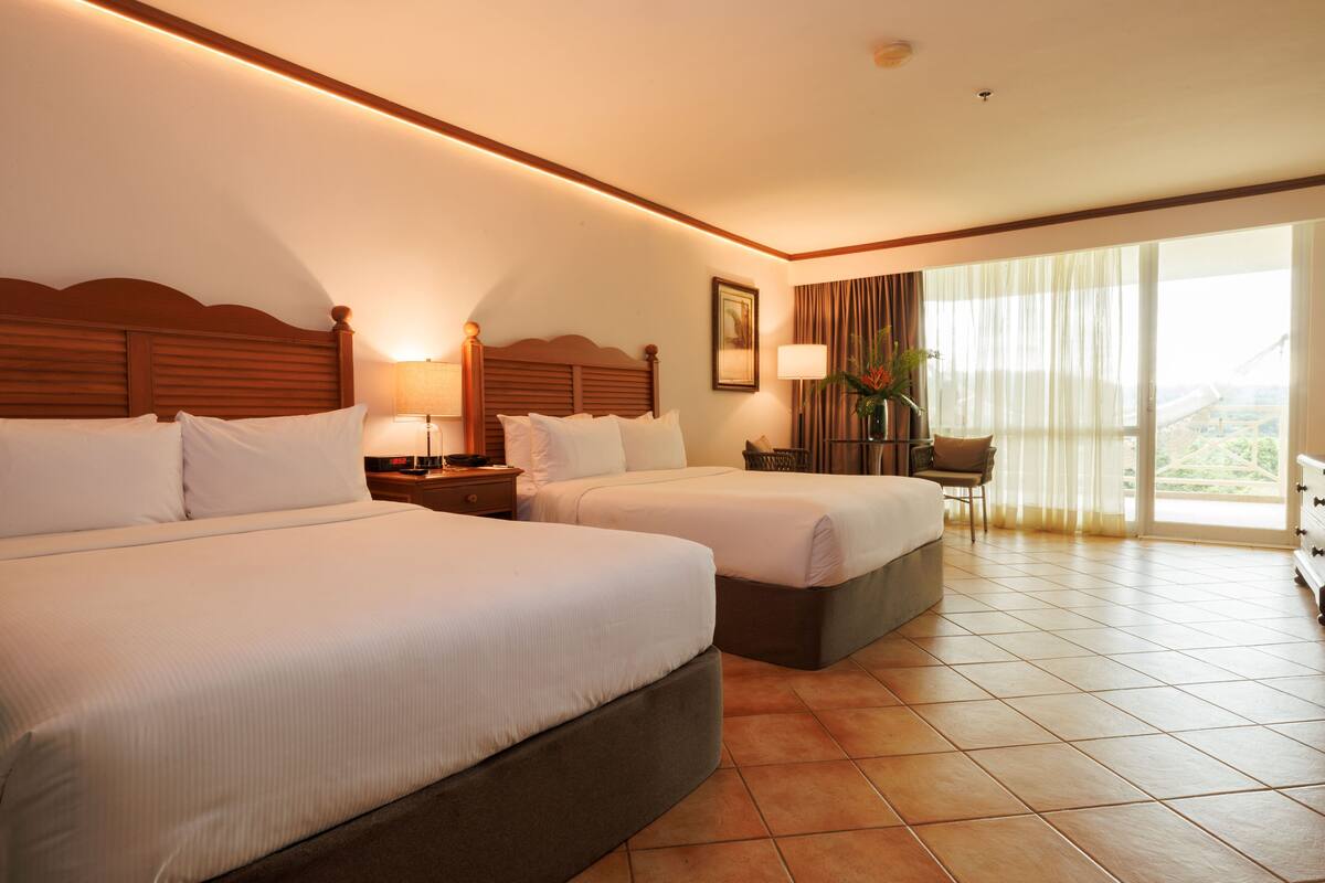 A hotel room with two beds, a desk, a chair, and a large window with curtains.