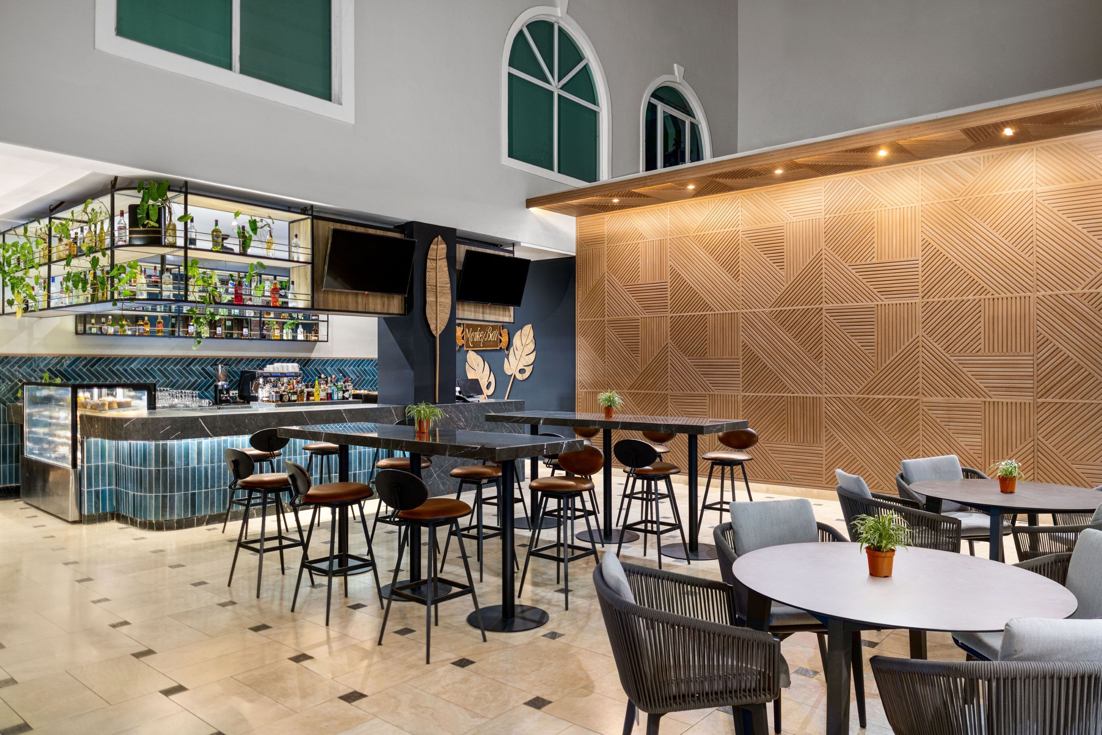 Un bar moderno con barra, taburetes y una variedad de botellas. Hay mesas y sillas para sentarse, y una pared decorativa con patrones geométricos.