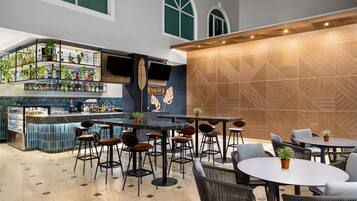 Un bar moderno con barra, taburetes y una variedad de botellas. Hay mesas y sillas para sentarse, y una pared decorativa con patrones geométricos.