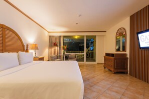 Quarto de hotel com cama grande, televisão, varanda com mesa e cadeiras, e cômoda de madeira.