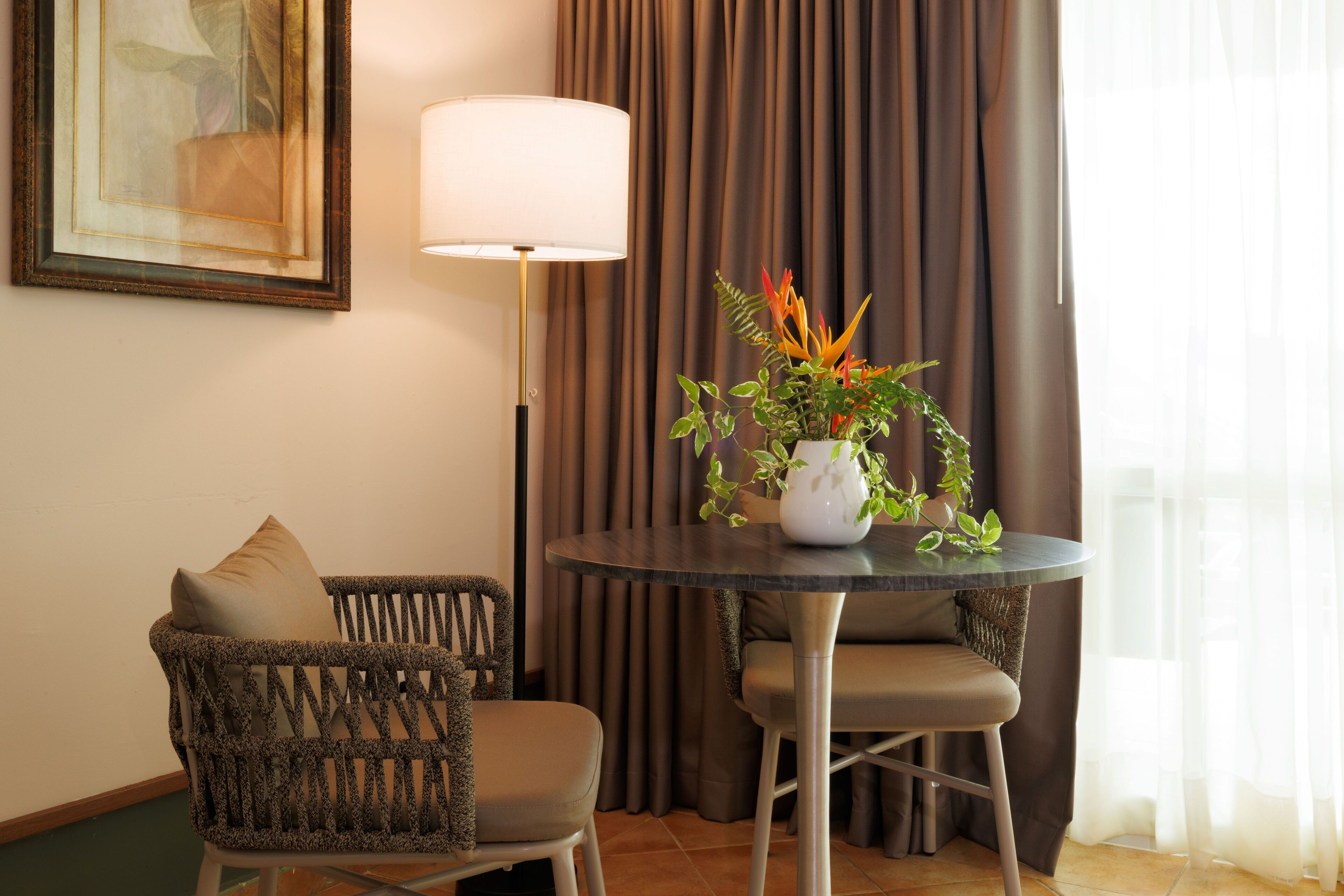 Una habitación con una mesa redonda, dos sillas de mimbre, una lámpara de pie, una obra de arte enmarco y una planta en maceta.