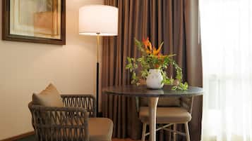 A room with a round table, two wicker chairs, a floor lamp, a framed artwork, and a potted plant.
