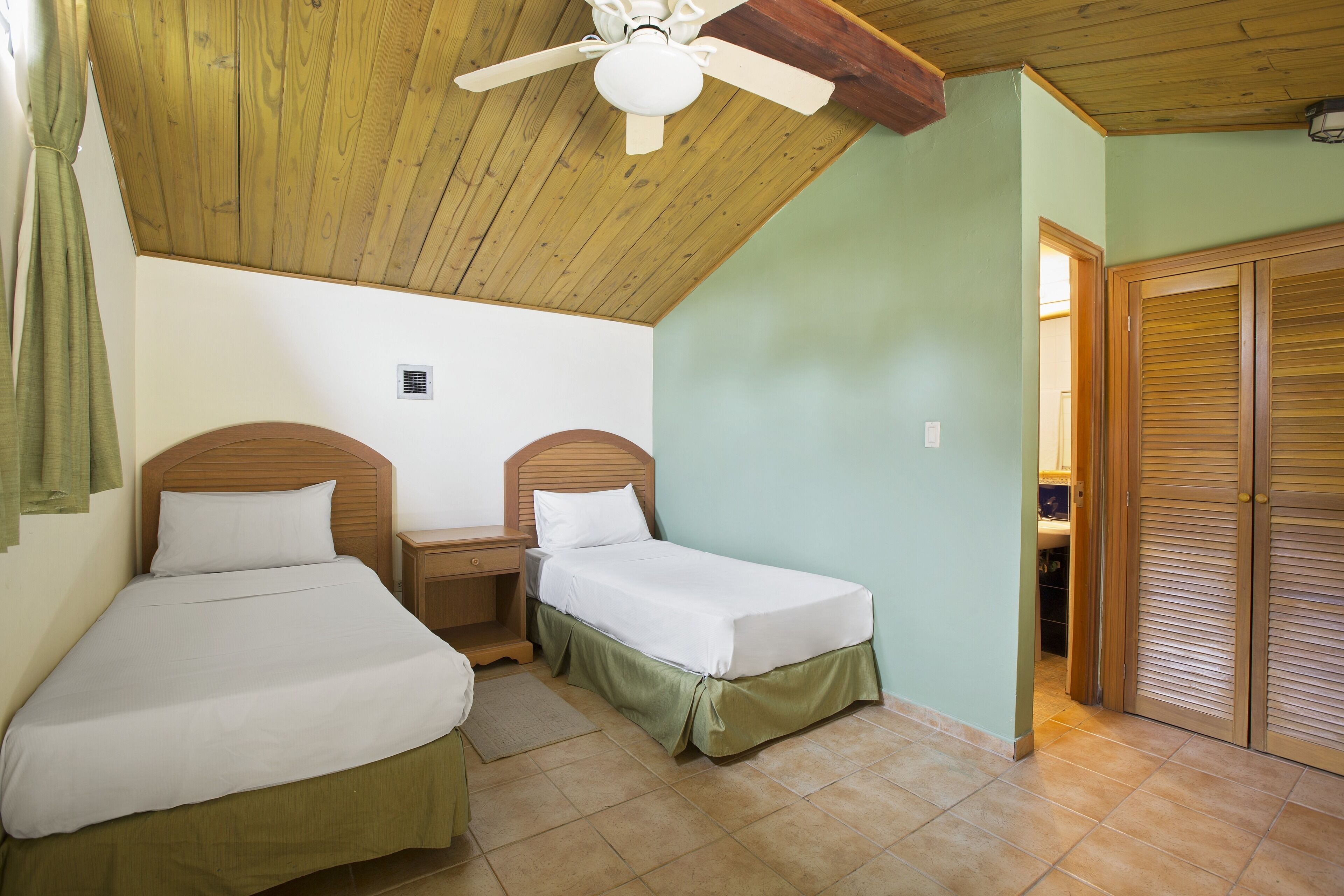 Une chambre avec deux lits simples, un ventilateur de plafond en bois et un mur vert.
