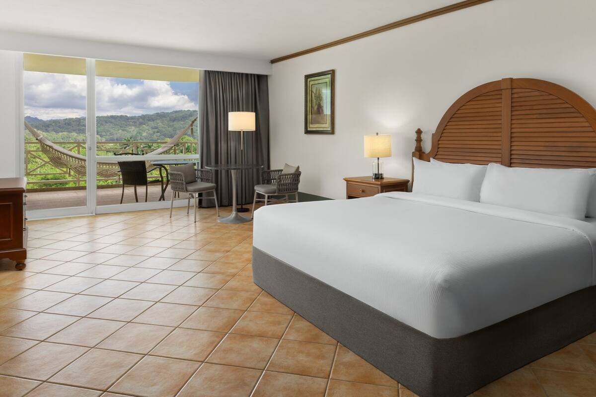 A hotel room with a large bed, a desk with chairs, a view of the outdoors, and a painting on the wall.