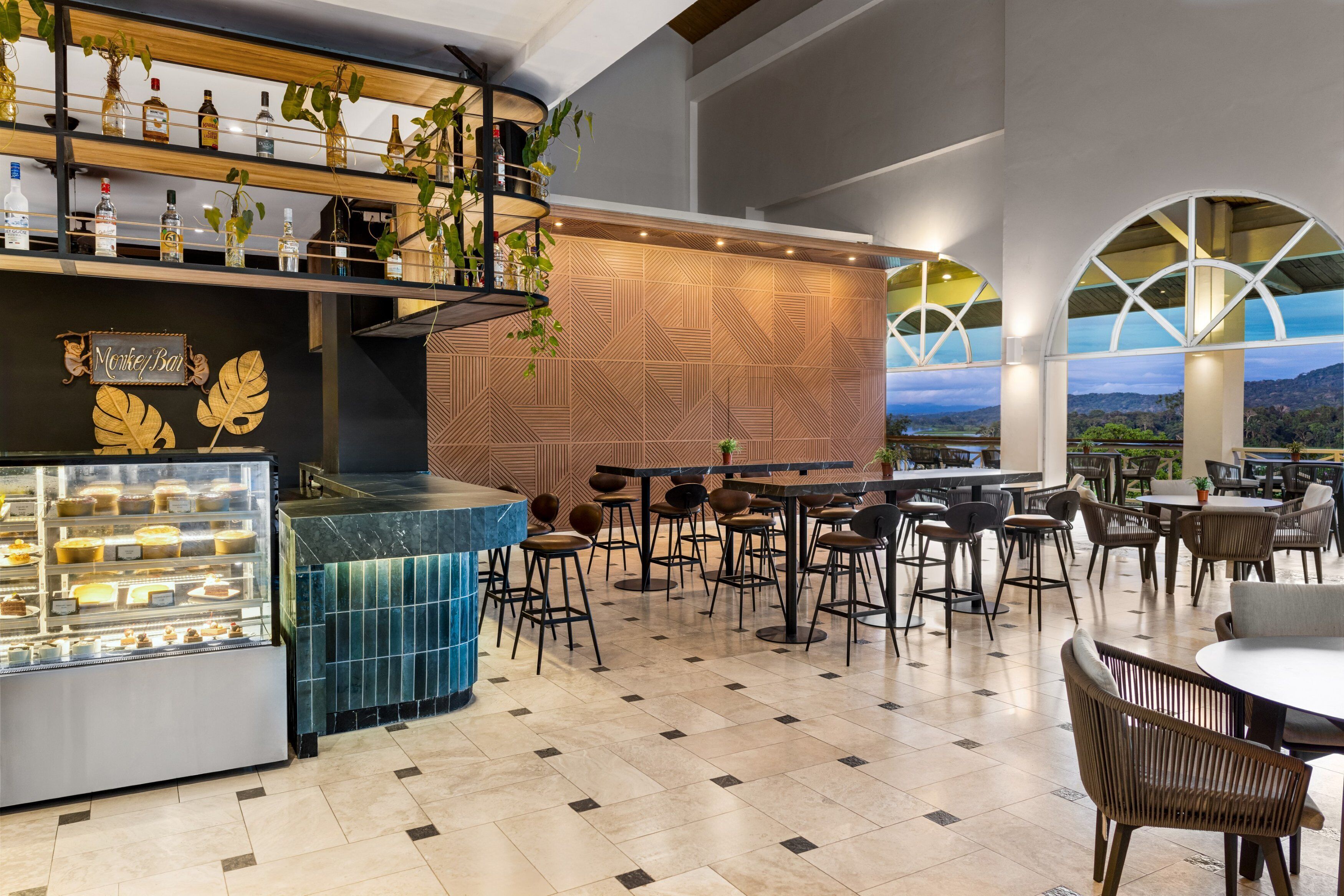 Uma área de bar moderna com um expositor de sobremesas, prateleiras de madeira com garrafas e um espaço de estar com mesas e cadeiras.