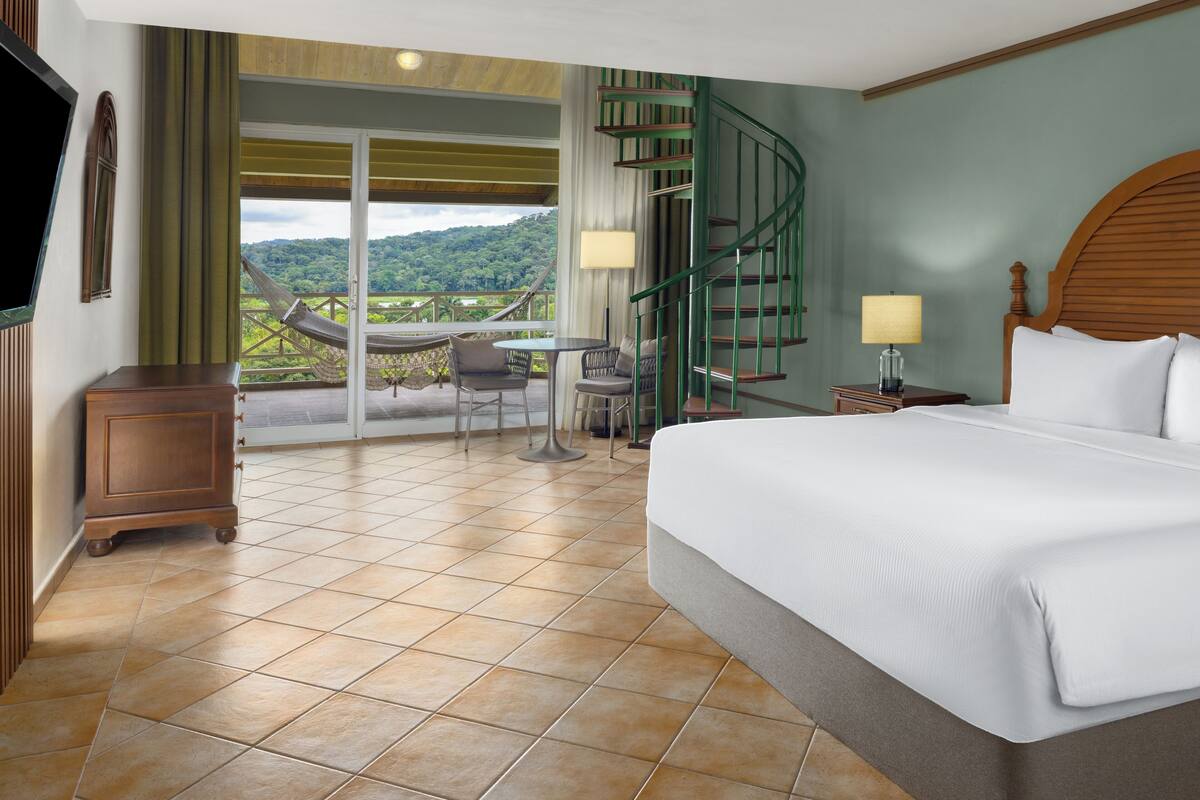 A hotel room with a bed, a TV, a balcony with a view, a table with chairs, and a spiral staircase.