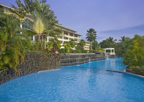 Piscina de resort com muro de pedra, palmeiras e um edifício com várias andares e varandas.
