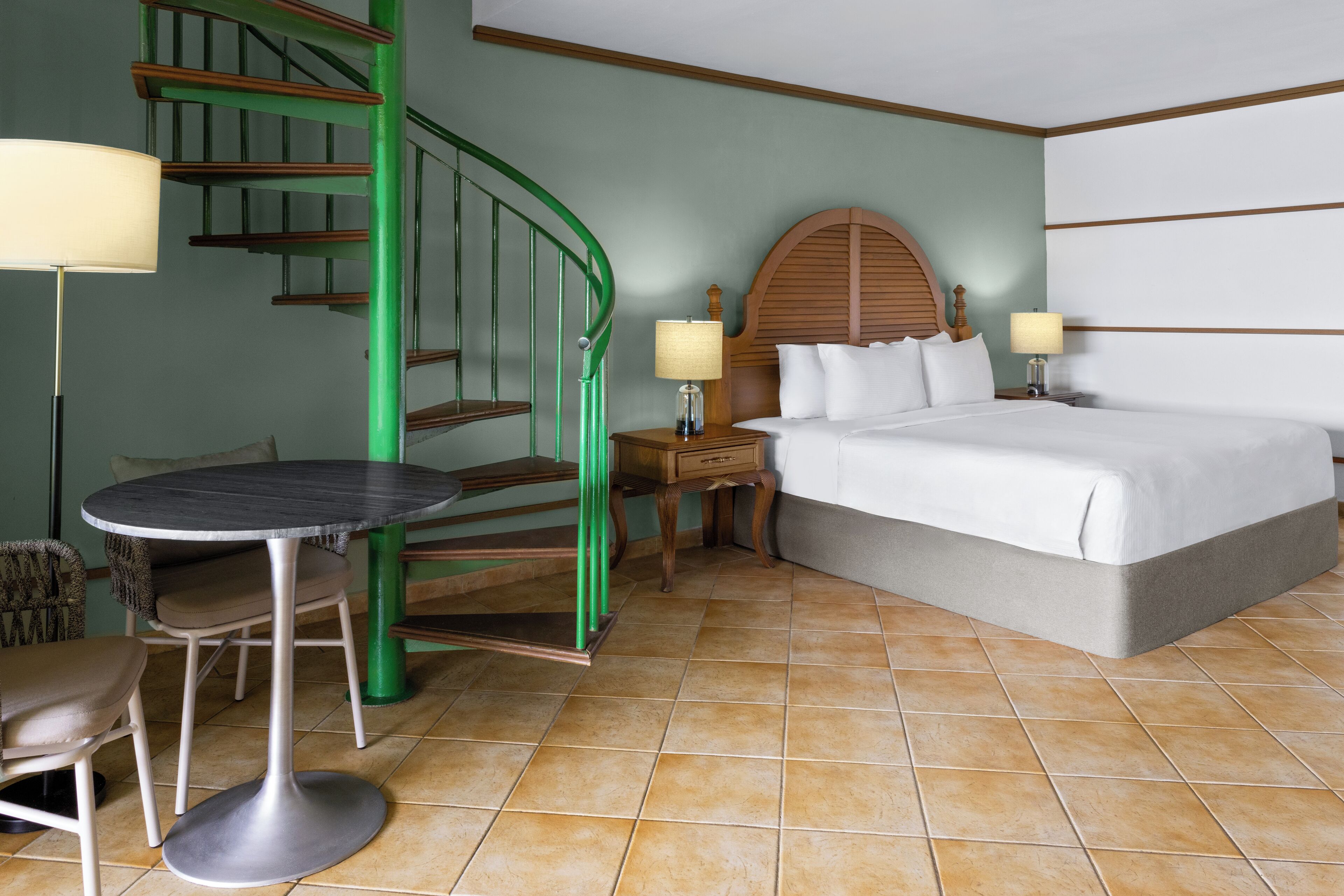 A hotel room with a bed, a round table, a chair, a nightstand, a lamp, and a green spiral staircase.