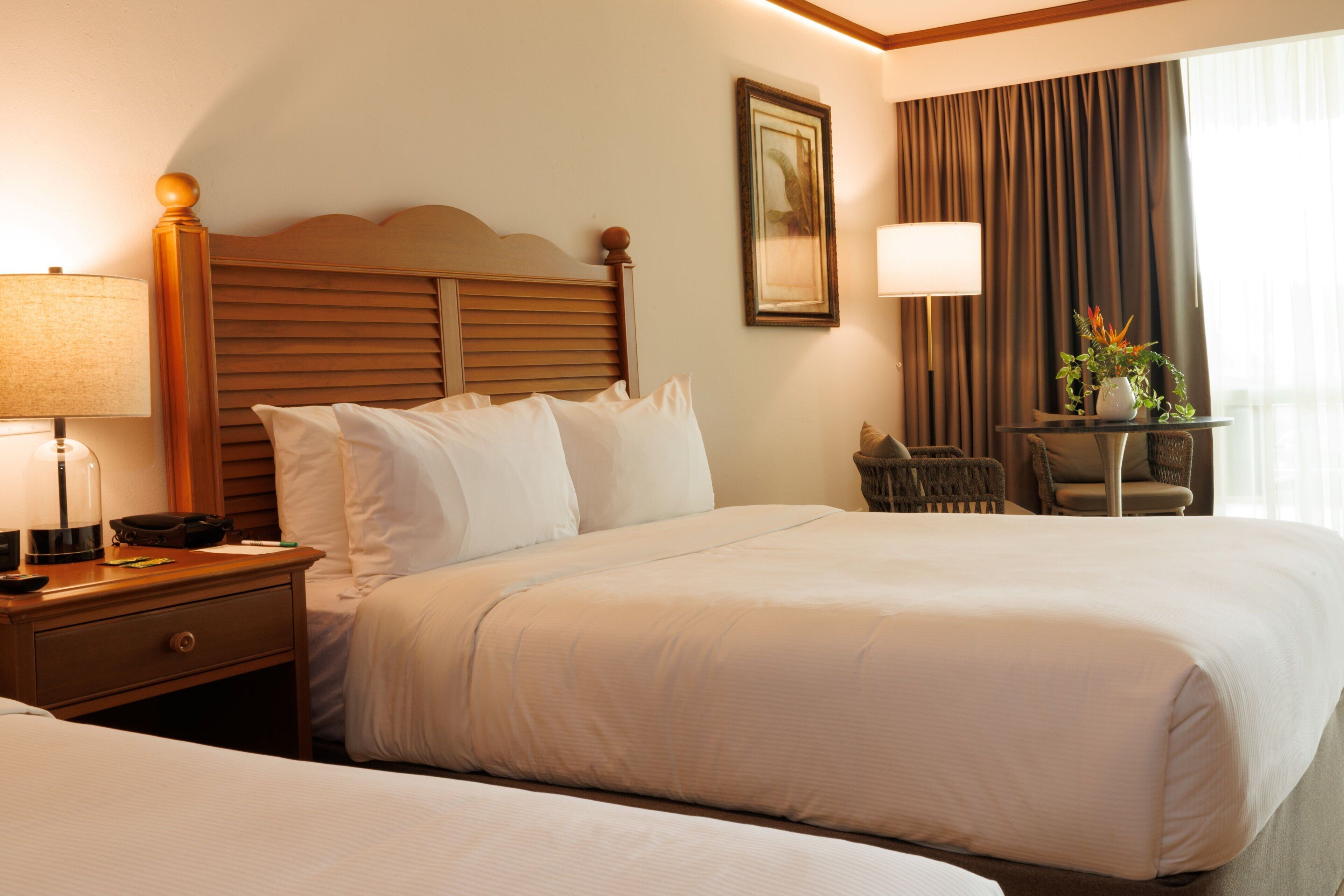 A hotel room with a large bed, a bedside table with a lamp, a chair, and a small table with a plant.