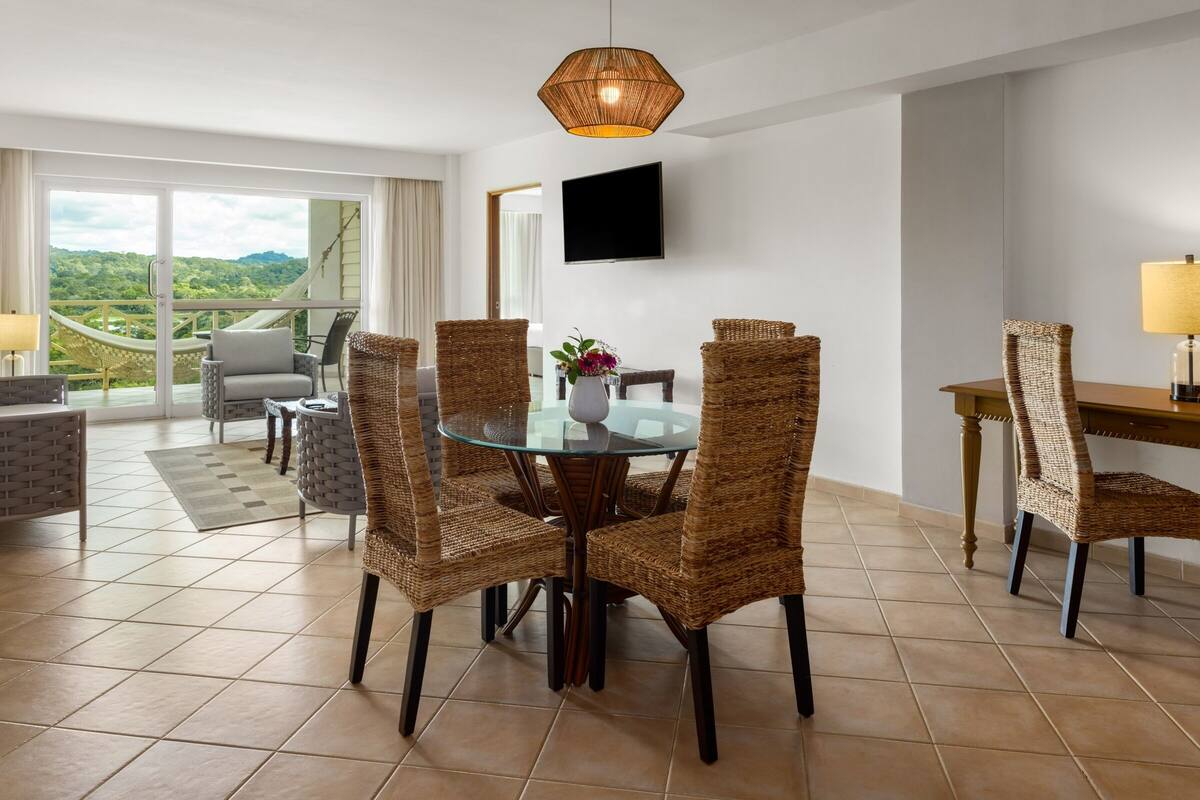 A dining area with wicker chairs, a glass table, and a view of the outdoors.