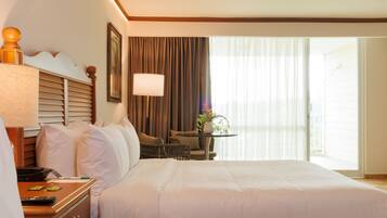 A hotel room with a large bed, a bedside table with a lamp, a chair, a small table with a plant, and a view of the outdoors through the window.