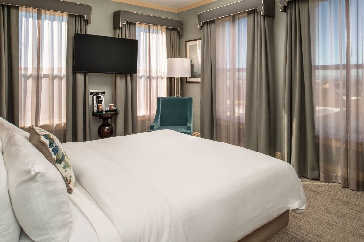 Habitación de hotel con una cama grande, un televisor de pantalla plana, una silla y una mesita con lámpara y cafetera.