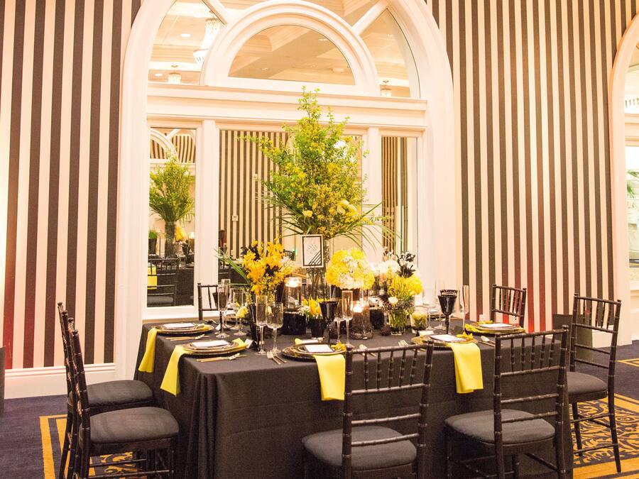 Ein formeller Speisesaal mit einem langen Tisch, der für eine Mahlzeit gedeckt ist, umrahmt von hohen Stühlen und einer großen Vase mit gelben Blumen in der Mitte.