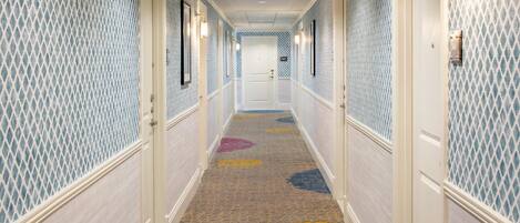 Un pasillo de hotel con papel tapiz estampado, una alfombra con diseños coloridos y una puerta al final.