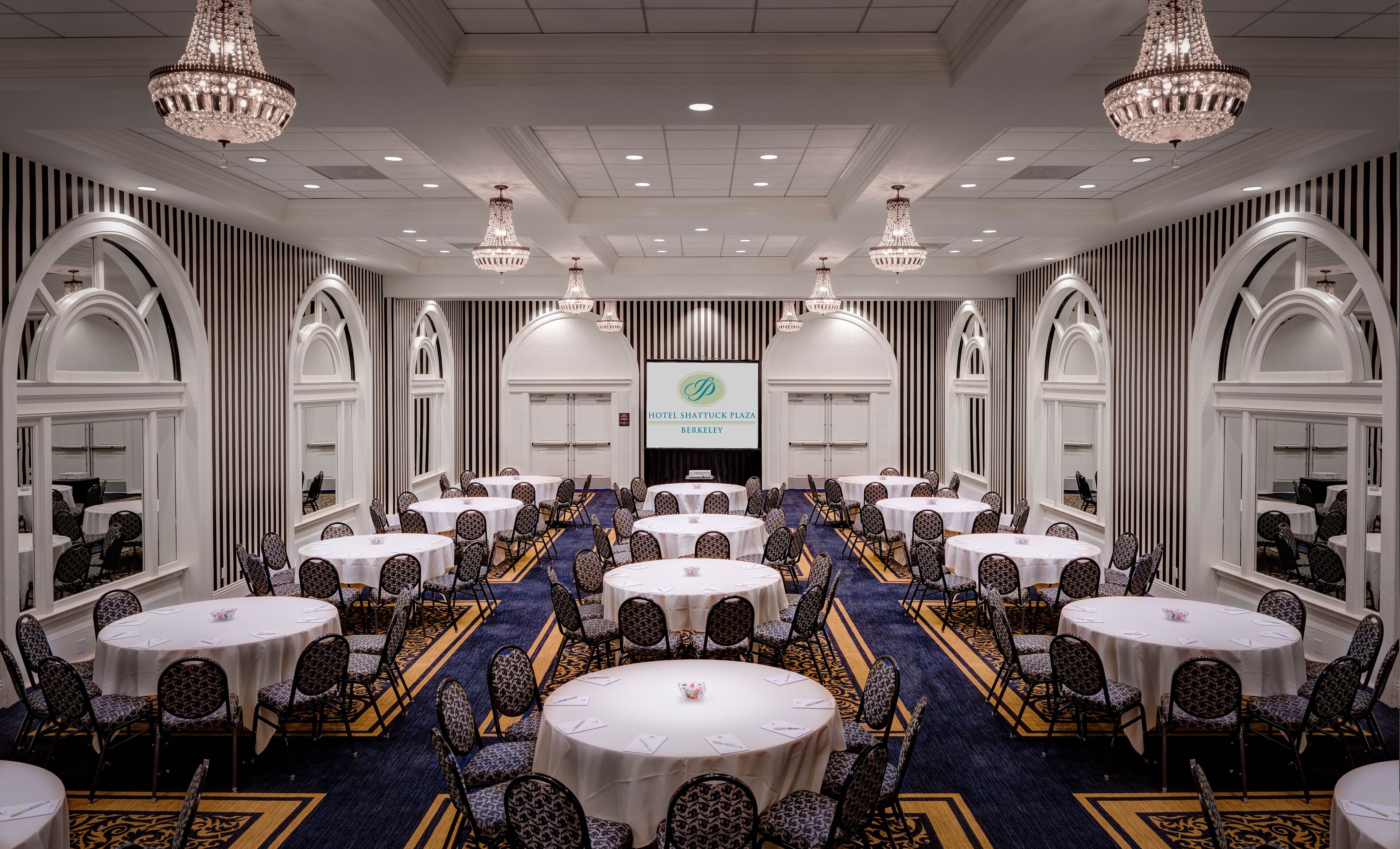 A large conference room with multiple round tables and chairs, chandeliers, and a screen displaying the hotel name.