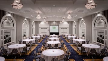 A large conference room with multiple round tables and chairs, chandeliers, and a screen displaying the hotel name.