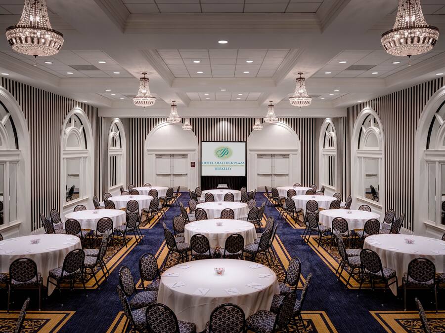 A large conference room with multiple round tables and chairs, chandeliers, and a screen displaying the hotel name.