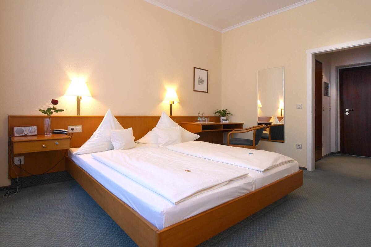 A hotel room with a double bed, bedside tables, a desk, a chair, a lamp, a mirror, and a door.