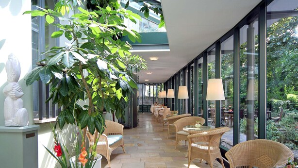 A sunlit corridor with wicker furniture, large windows, and a central plant. - Hotel Martha Dresden (Dresden)
