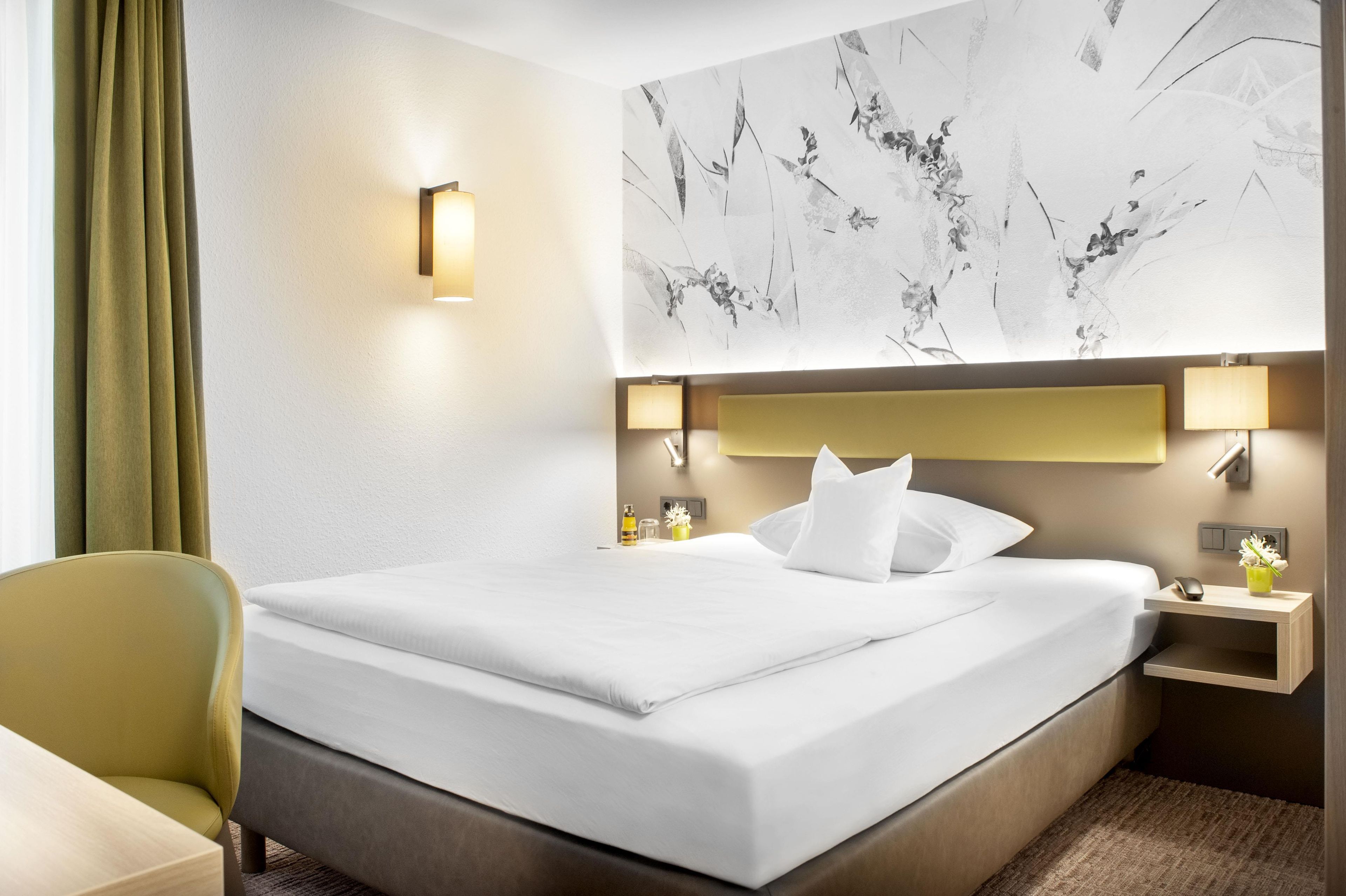 A hotel room with a large bed, two bedside lamps, a nightstand, a chair, and a wall with a floral mural.