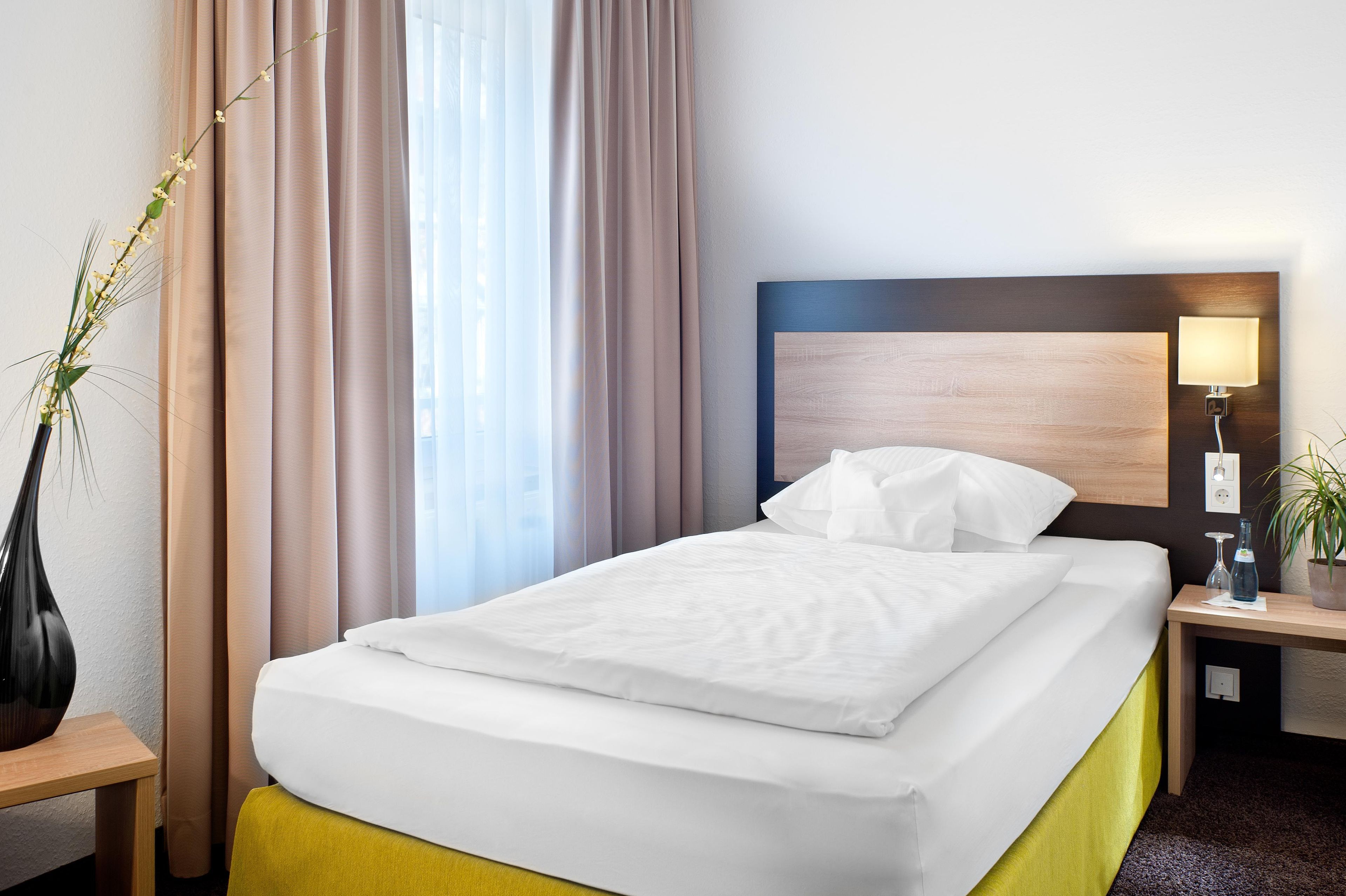 Une chambre d’hôtel avec un lit bien fait, une table de chevet avec une lampe, une plante en pot et une fenêtre avec des rideaux.
