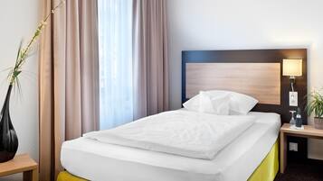 Une chambre d’hôtel avec un lit bien fait, une table de chevet avec une lampe, une plante en pot et une fenêtre avec des rideaux.