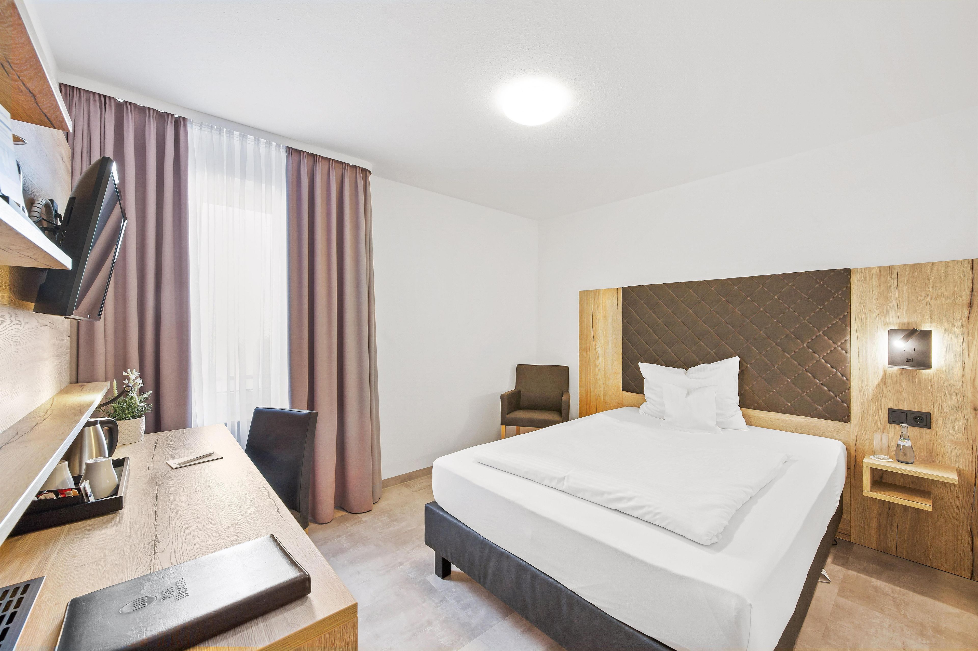 Una camera d'hotel con un letto, una scrivania con un portatile, una sedia, una TV e un comodino con una bottiglia.