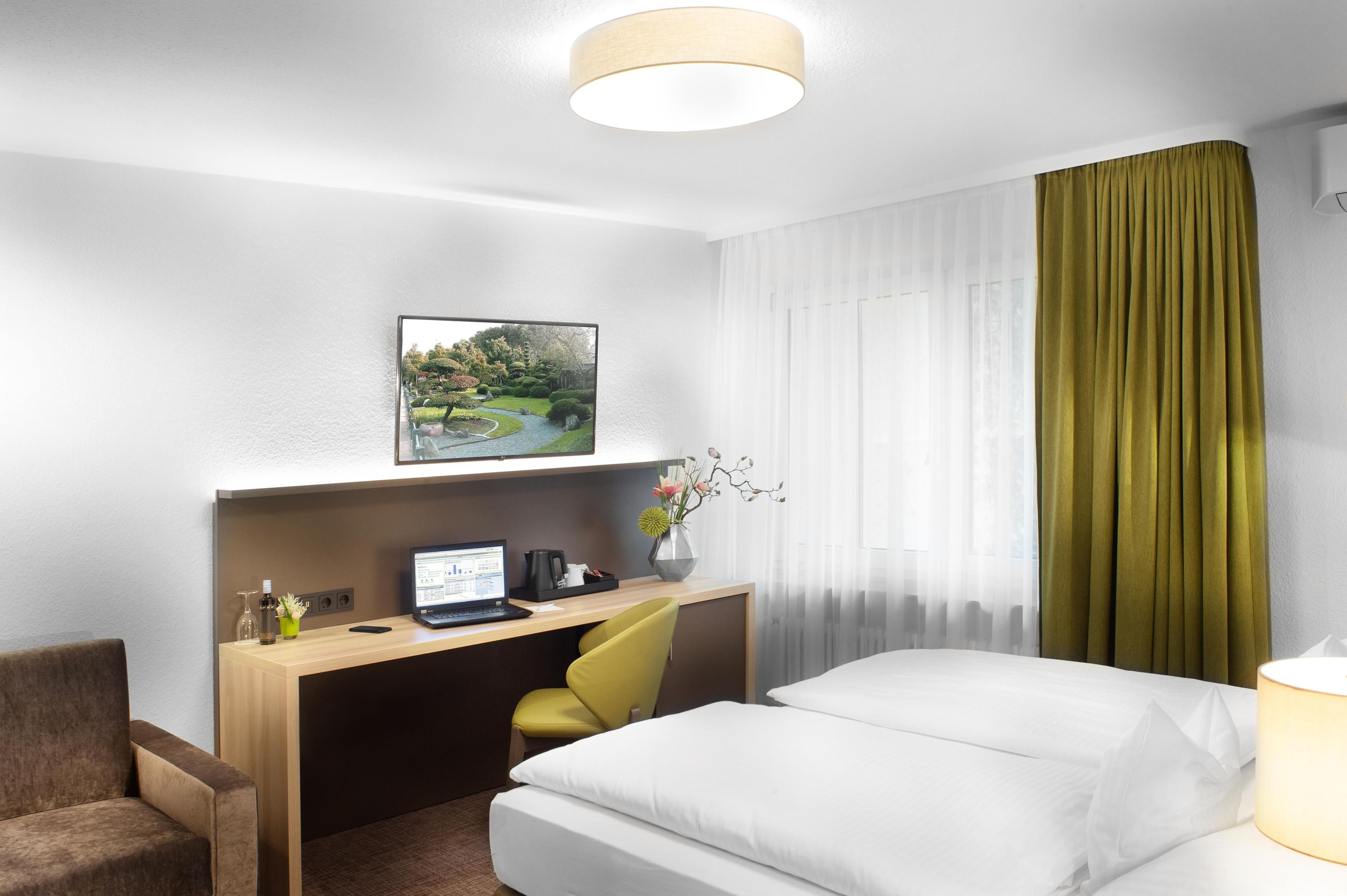 A hotel room with a bed, a desk with a laptop, a chair, a small table with a vase, and a painting on the wall.