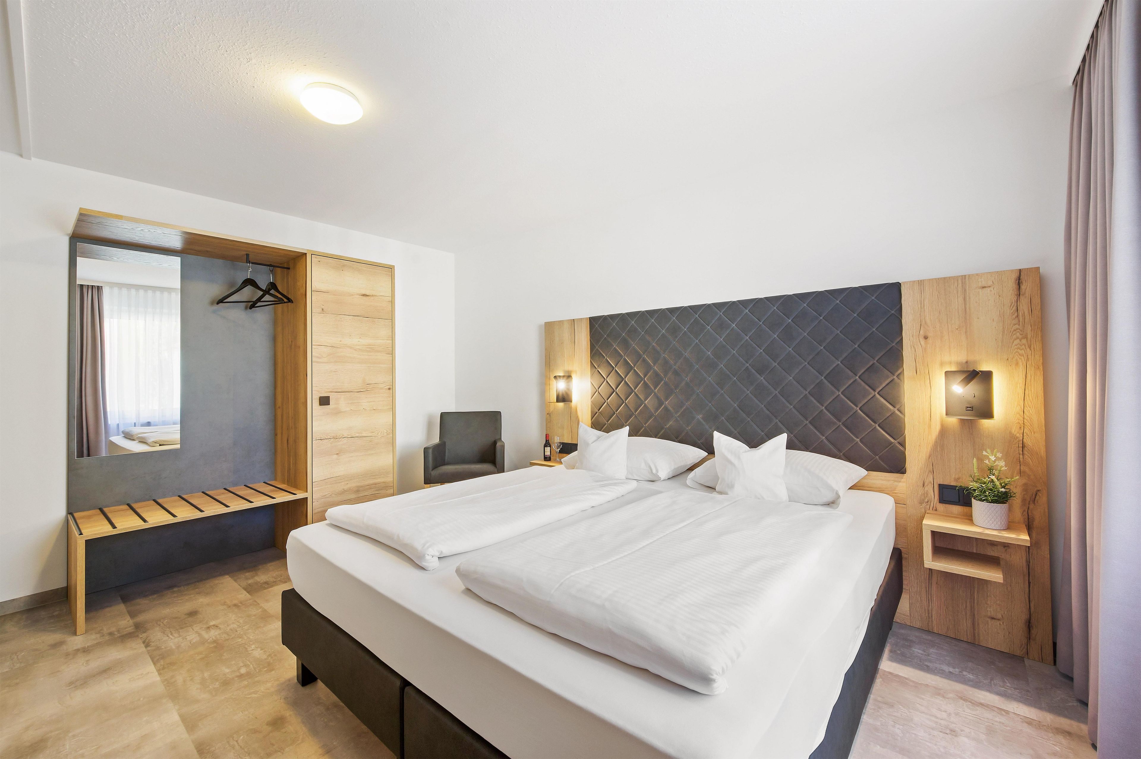 A modern hotel room with a large bed, a wooden bench, a mirror, and a wardrobe.