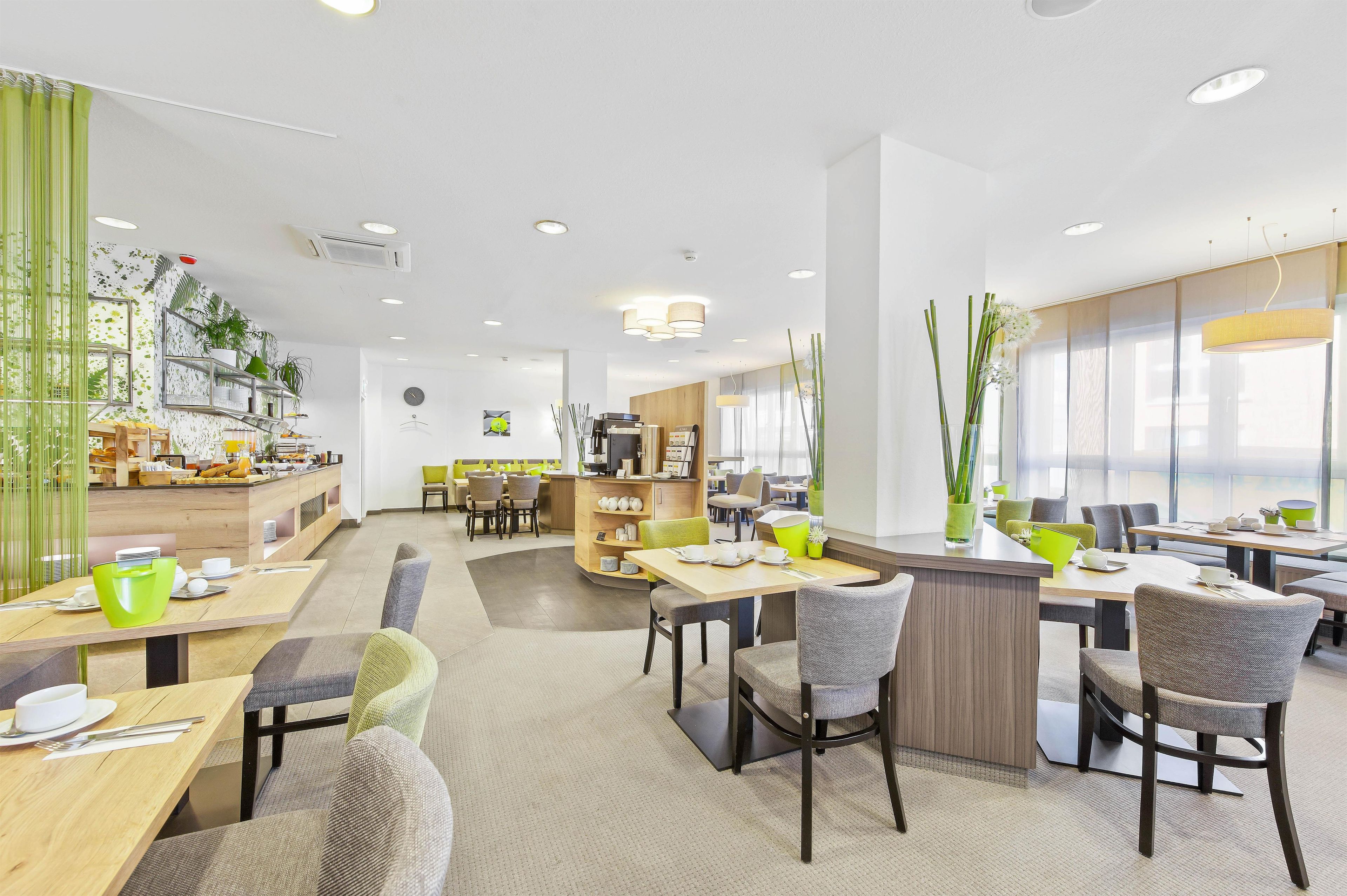 A modern dining area with multiple tables and chairs, a kitchen area in the background, and large windows allowing natural light.