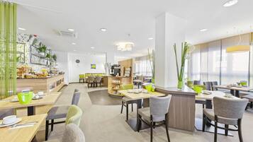 A modern dining area with multiple tables and chairs, a kitchen area in the background, and large windows allowing natural light.