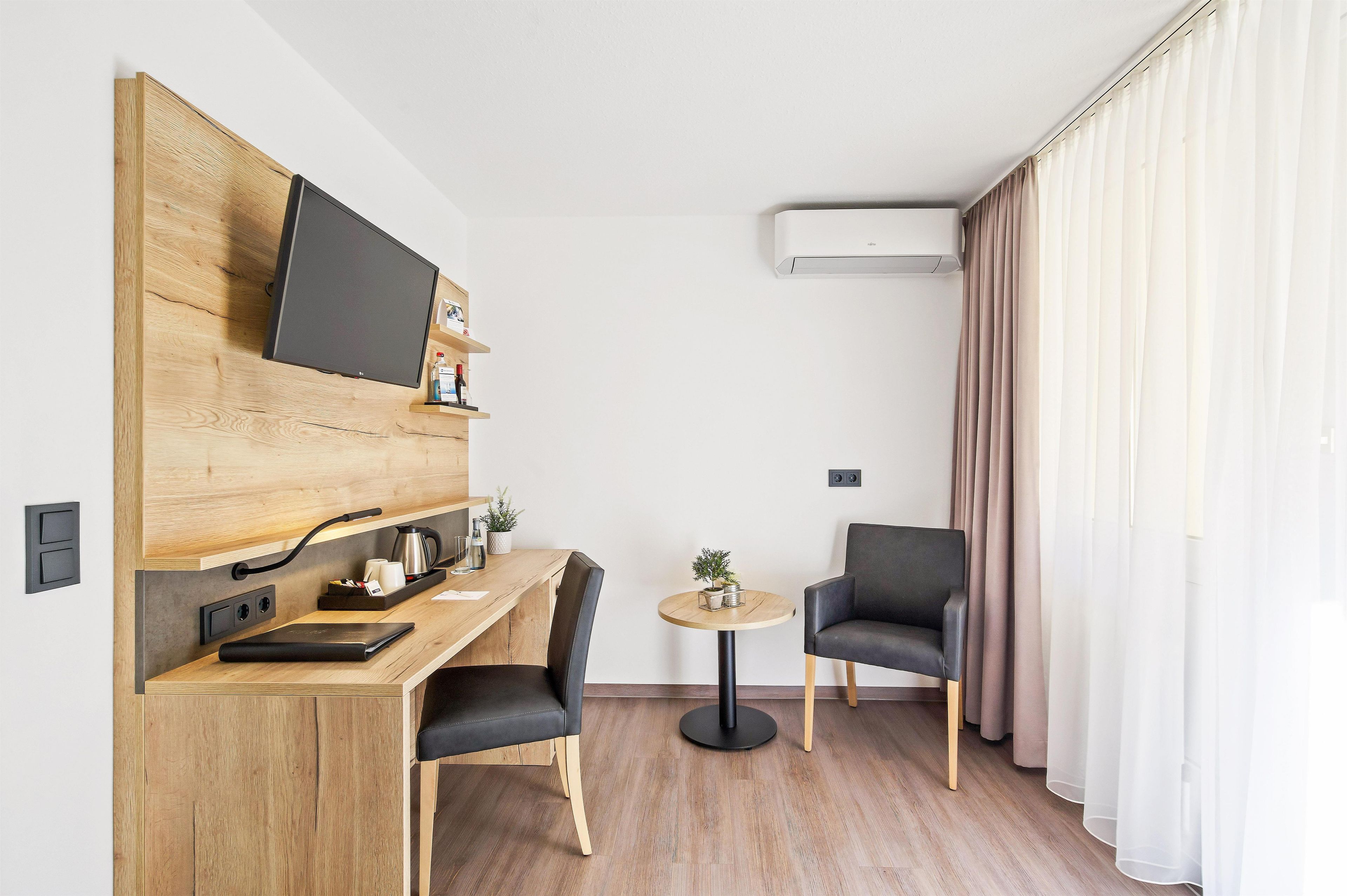 Moderni hotellihuone, jossa on puinen työpöytä, taulutelevisio, pieni pyöreä pöytä, kaksi tuolia ja suuri ikkuna, jossa on verhot.