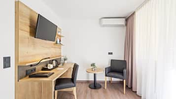 Moderni hotellihuone, jossa on puinen työpöytä, taulutelevisio, pieni pyöreä pöytä, kaksi tuolia ja suuri ikkuna, jossa on verhot.