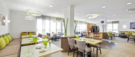 A modern dining area with multiple tables and chairs, a central bar area, and large windows allowing natural light.