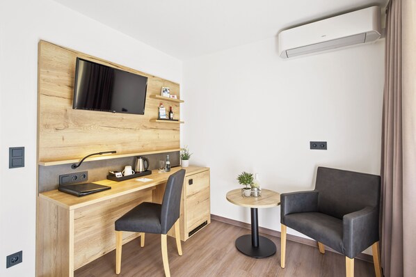 Ein kleines Hotelzimmer mit einem Holztisch, einem schwarzen Stuhl, einem runden Tisch mit Pflanze und einer schwarzen Couch.