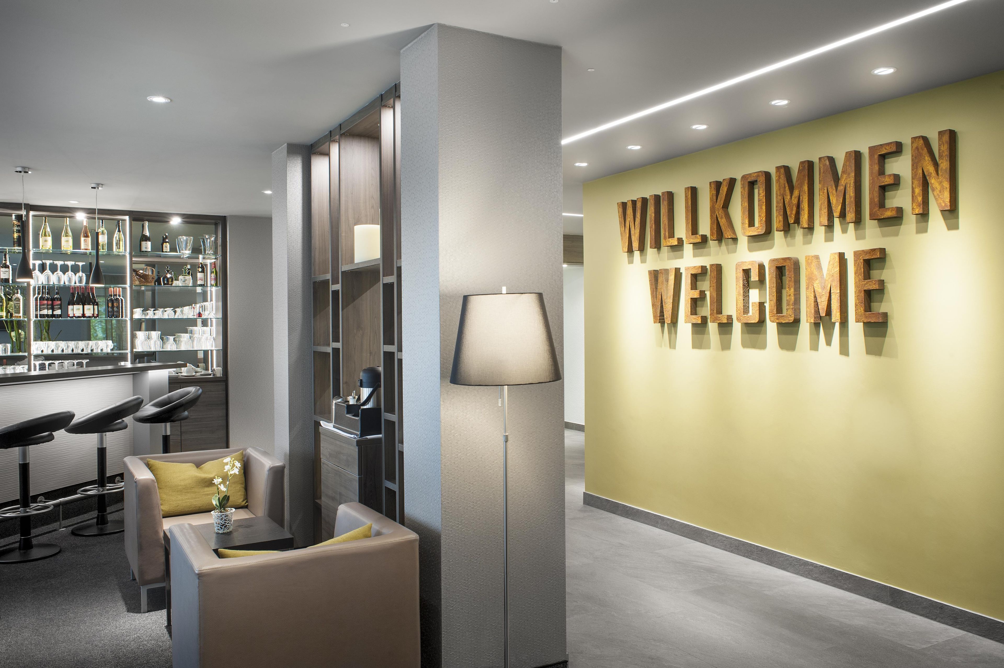 A modern reception area with a bar, seating, and a wall displaying 'WILKOMMEN WELCOME'.