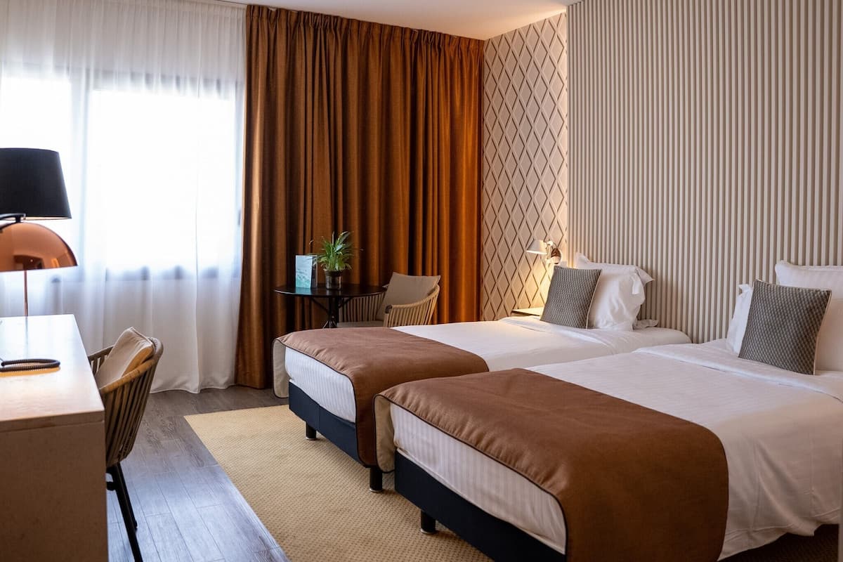 Une chambre d’hôtel avec deux lits, un bureau, une chaise, une lampe et une fenêtre avec des rideaux.